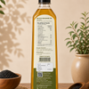 A bottle of black sesame oil stands upright on a wooden surface, showing the back label with nutrition facts and product details. The bottle is surrounded by loose black sesame seeds in wooden bowls and a spoon, with potted plants and leaf shadows in the background.
