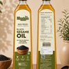 Two bottles of Meadow's Black Sesame Oil displayed on a wooden surface, showing both the front label and the back nutrition panel. The bottles are filled with golden oil and are framed by scattered sesame seeds, a small wooden bowl, and soft-focus green plants against a warm beige background.