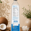 A tall clear bottle of coconut oil with a black cap stands on a wooden surface, showing the back label with nutrition facts and product details. The scene is styled with a halved coconut, a small bowl of white flakes, and potted plants against a warm beige background.