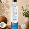 A bottle of coconut oil stands upright on a wooden surface, showing the back label with nutrition facts and product details. The bottle is styled with a whole coconut, a cut coconut, a small bowl of white coconut pieces, and potted plants against a warm beige background.