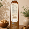 A tall bottle of groundnut oil shown from the back, with a black cap and a beige nutrition label. The bottle is placed on a wooden surface with peanuts in a bowl and scoop, and potted plants in the background.