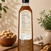 A bottle of groundnut oil stands on a wooden table with peanuts in a bowl and scattered peanuts around it, set against a warm beige background with potted plants.
