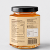 Back view of a clear glass jar of honey with a black lid and a beige label showing nutritional information, marketing details, and a barcode, on a light gray background.