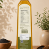 A bottle of black sesame oil stands upright on a wooden surface, showing the back label with nutrition facts and product details. The bottle is surrounded by loose black sesame seeds in wooden bowls and a potted plant, with a warm beige background and soft leaf shadows.