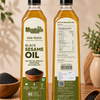 Two bottles of Meadow's Black Sesame Oil shown side by side on a wooden surface, displaying both the front label and the back nutrition panel. The bottles are filled with golden-yellow oil and are surrounded by loose black sesame seeds, bowls, and a warm, neutral background with soft-focus plants.